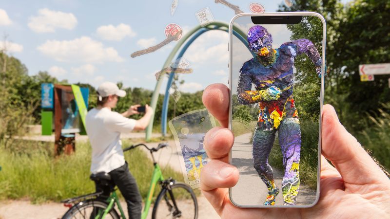 Spielplatz; davor Hand mit Smartphone, auf dessen Bildschirm virtuelle Zusatzinhalte zu sehen sind.