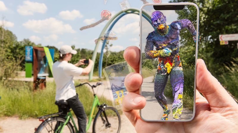 Spielplatz; davor Hand mit Smartphone, auf dessen Bildschirm virtuelle Zusatzinhalte zu sehen sind
