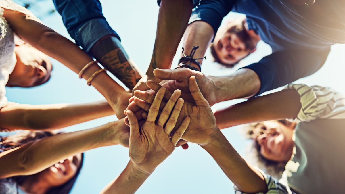Gruppe verschiedener Menschen legt gemeinsam die Hände übereinander – Symbol für Teamarbeit, Zusammenhalt und Gemeinschaft.