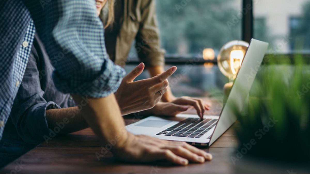 Keyvisual der IN.NRW: Zwei Menschen stehen an einem Holztisch und beugen sich über einen Laptop.