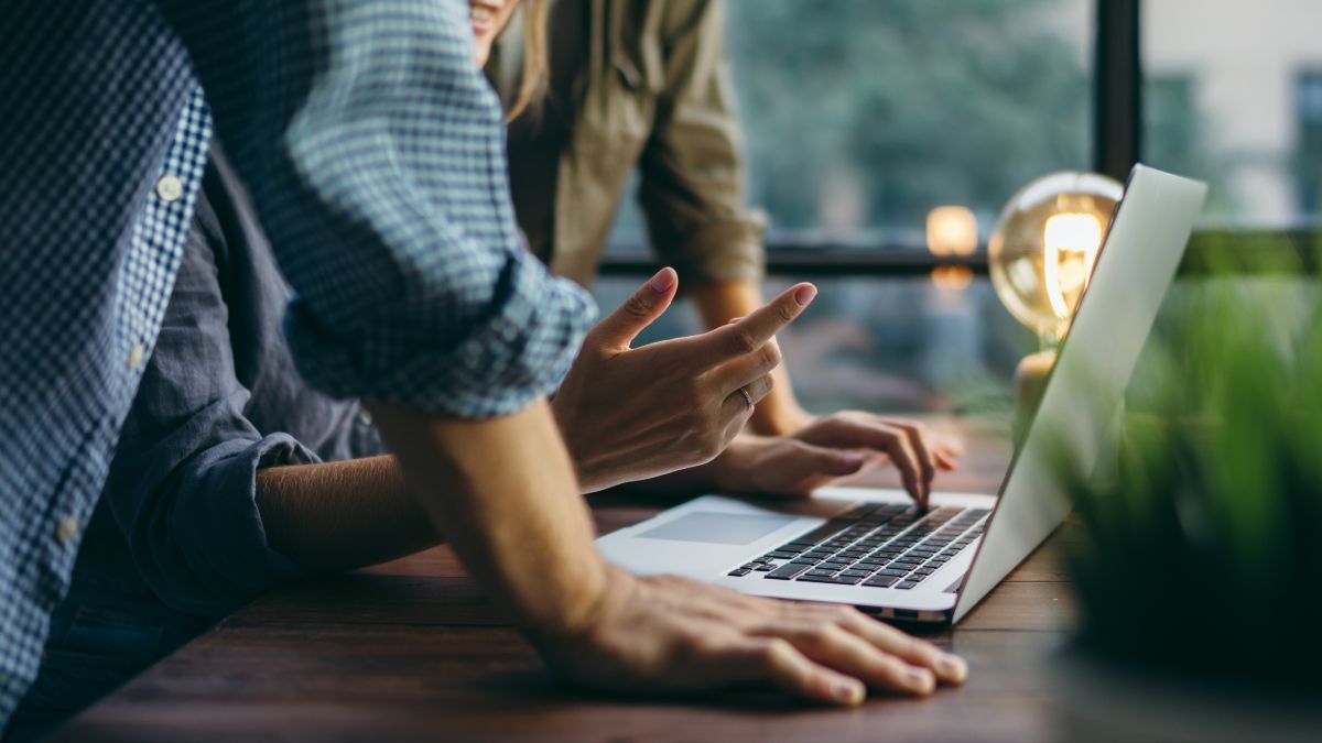  Keyvisual der IN.NRW: Zwei Menschen stehen an einem Holztisch und beugen sich über einen Laptop.