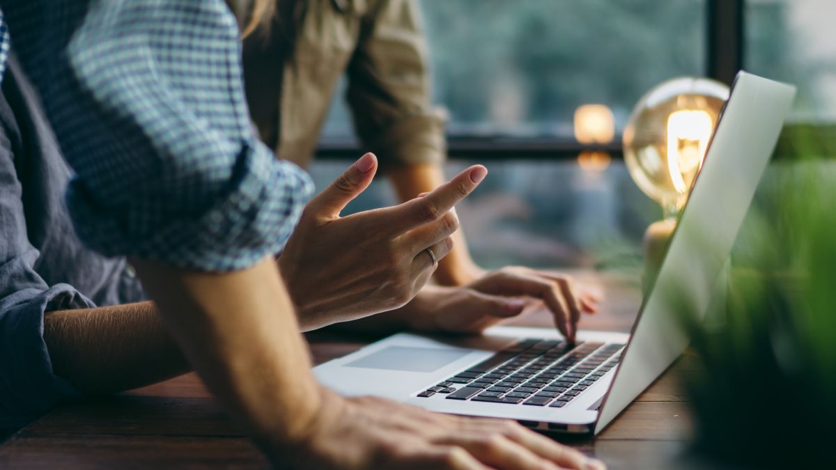 Keyvisual der IN.NRW: Zwei Menschen stehen an einem Holztisch und beugen sich über einen Laptop.