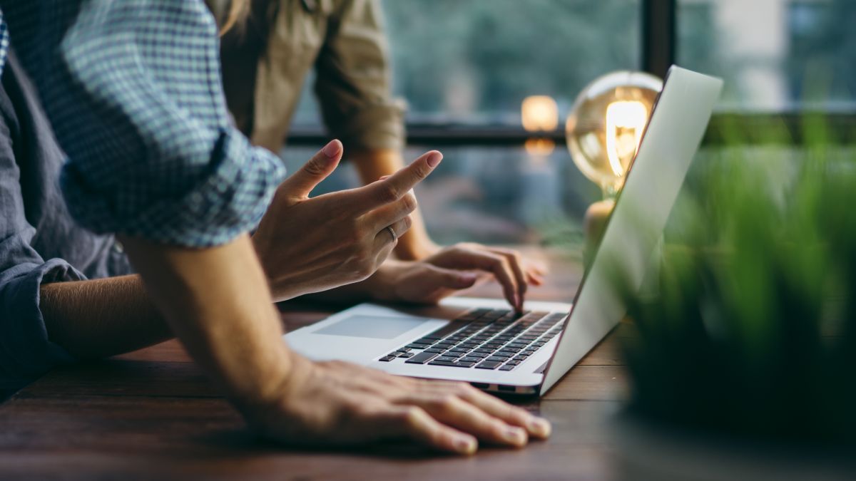 Keyvisual der IN.NRW: Zwei Menschen stehen an einem Holztisch und beugen sich über einen Laptop.