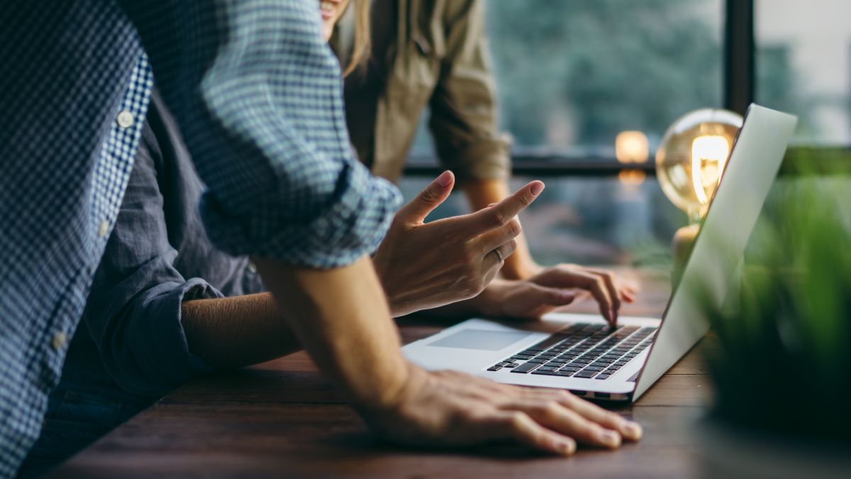 Keyvisual der IN.NRW: Zwei Menschen stehen an einem Holztisch und beugen sich über einen Laptop.