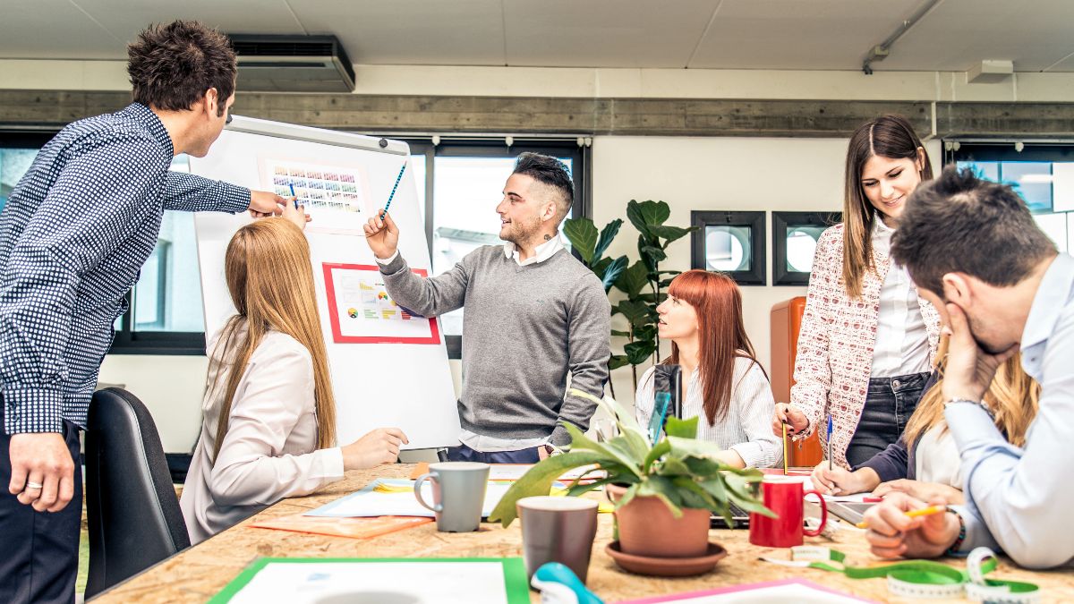 Fünf Personen, die an einem Tisch sitzen und/oder an einem Flipchart etwas zeigen