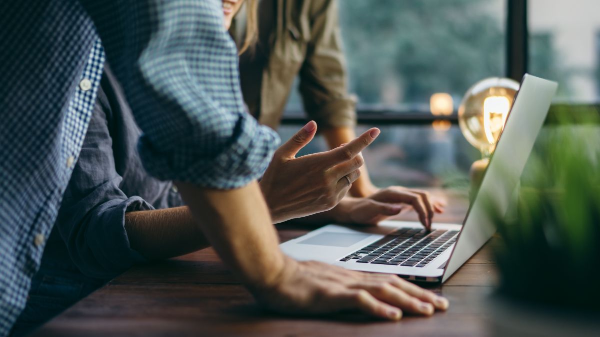 Keyvisual der IN.NRW: Zwei Menschen stehen an einem Holztisch und beugen sich über einen Laptop.